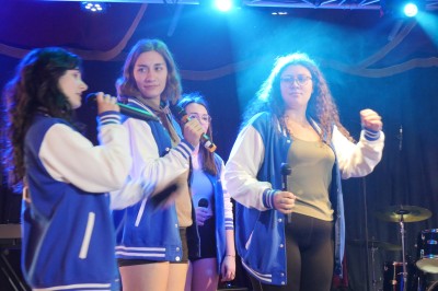 Des enfants colorient des dessins autour d'une table ronde, entourés d'adultes dans un espace intérieur animé.