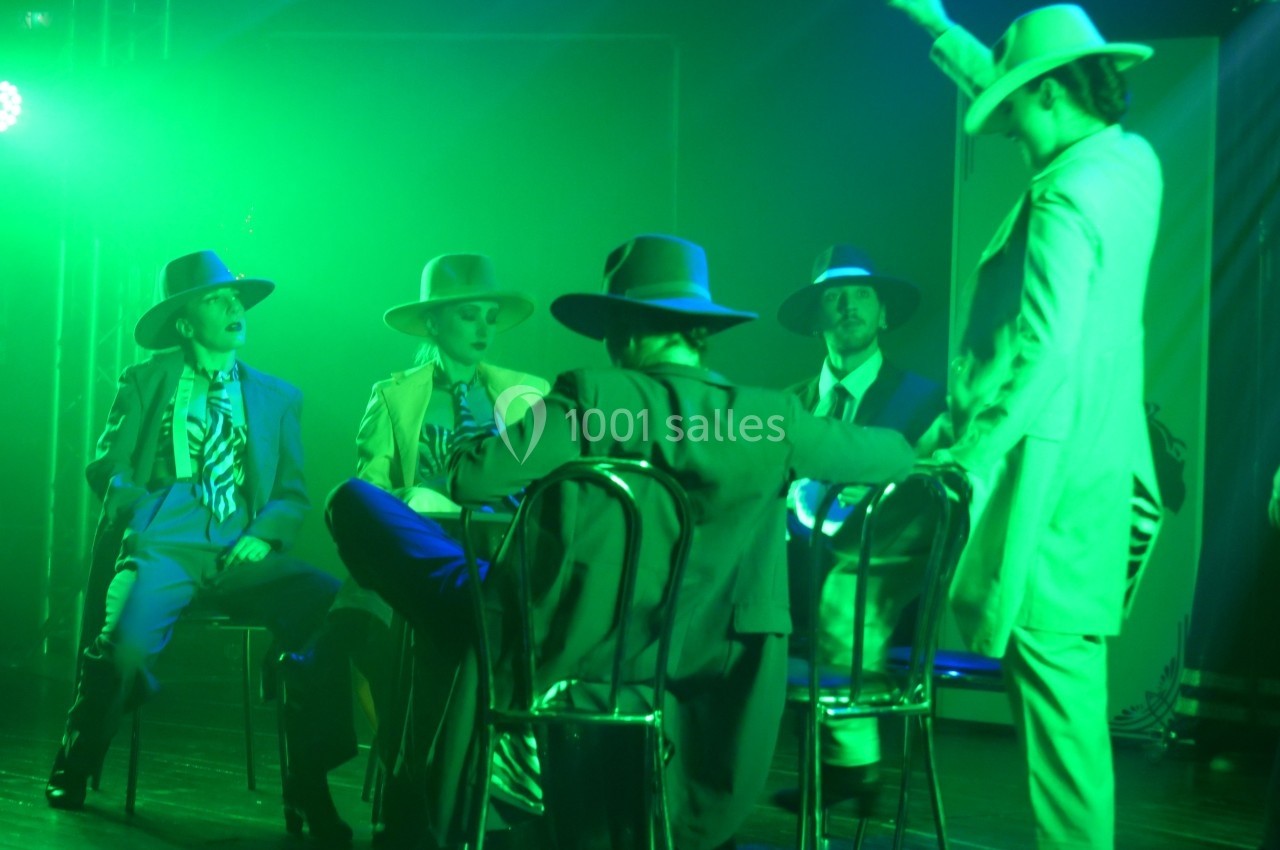 Des personnes en costumes et chapeaux assises sur des chaises, éclairées par une lumière verte dans une ambiance scénique.