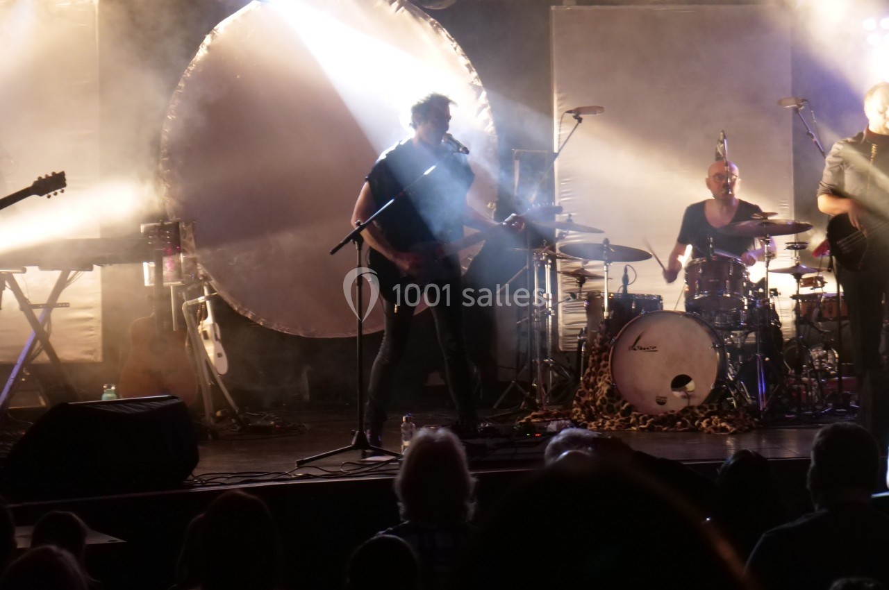 Un groupe joue sur scène dans une ambiance lumineuse tamisée, avec un batteur, un guitariste et des spectateurs en ombre.