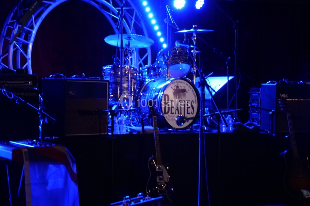 Batterie et guitares sur une scène éclairée, avec un logo ’Beatles’ visible sur la grosse caisse.