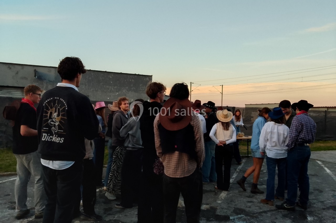 Un groupe de personnes, certaines portant des chapeaux, discute en extérieur sur un parking au coucher du soleil.