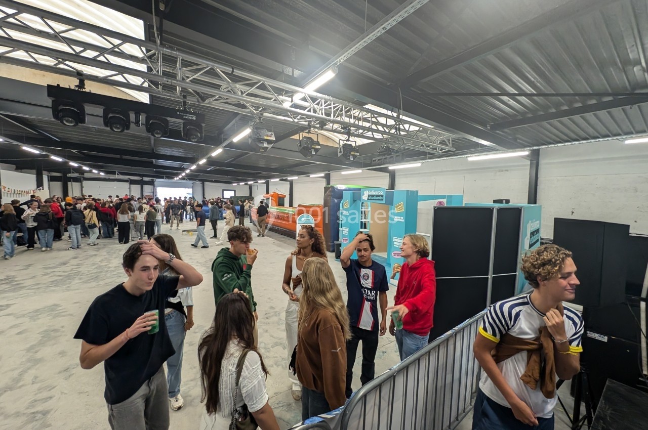 Des personnes discutent et se regroupent dans un grand espace intérieur avec des stands et une file d'attente au fond.