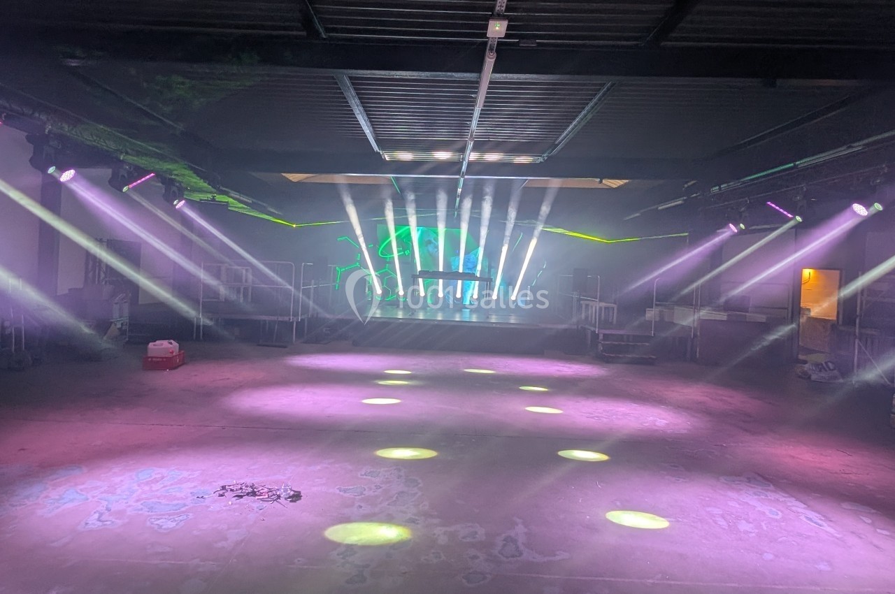 Salle vide avec éclairage coloré et faisceaux lumineux dirigés vers une scène au fond, dans un espace industriel.