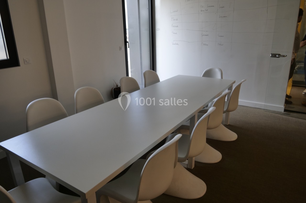 Salle de réunion avec une table blanche rectangulaire entourée de chaises modernes, devant un tableau blanc.