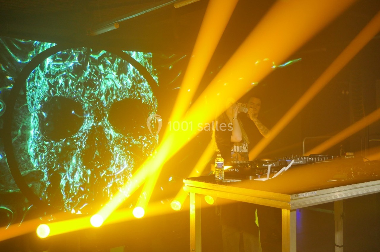 Un DJ mixe sur scène avec des effets lumineux jaunes et un écran affichant un crâne lumineux en arrière-plan.