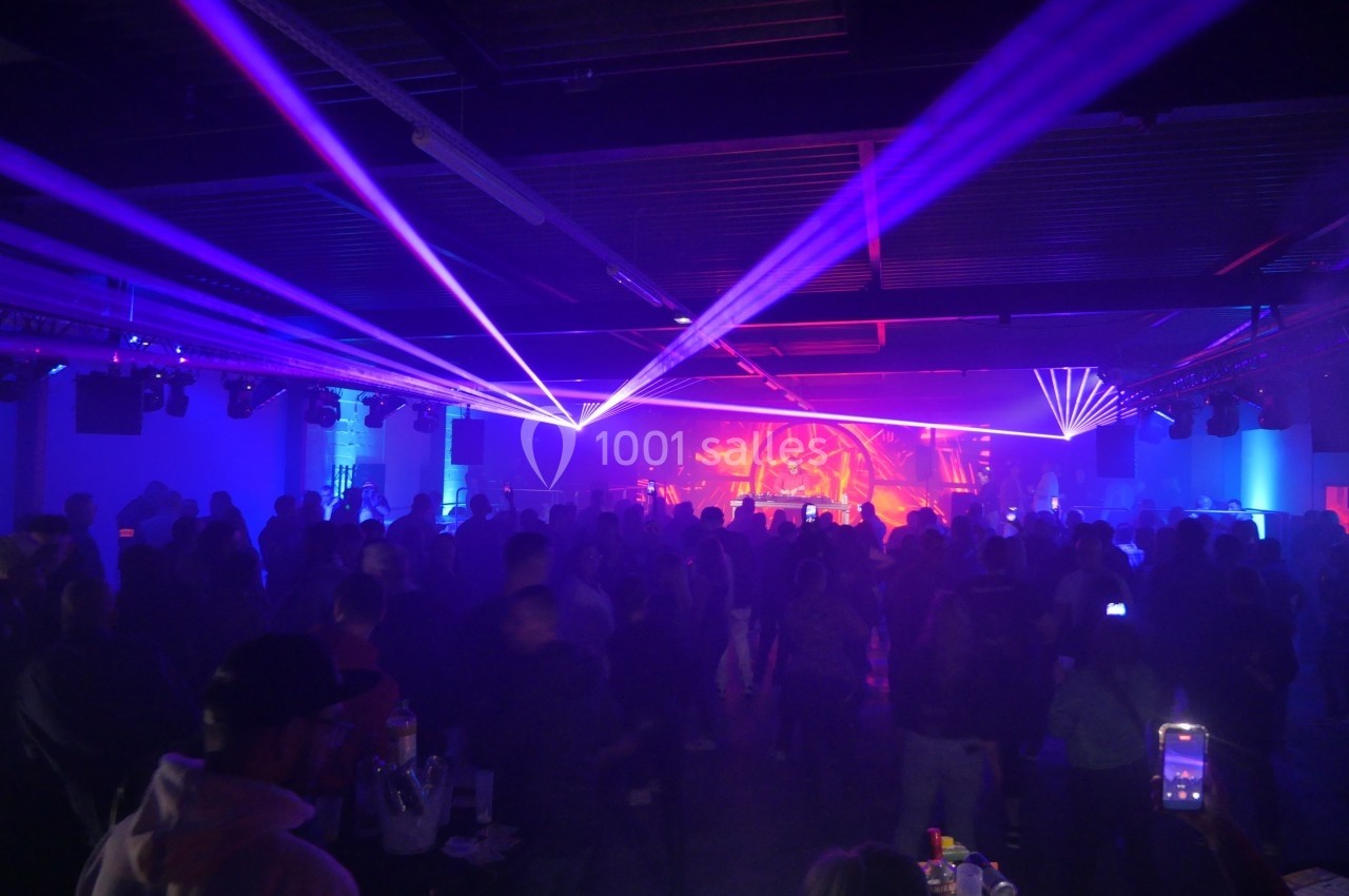 Salle sombre avec une foule debout, éclairée par des faisceaux lumineux colorés et une scène en arrière-plan.