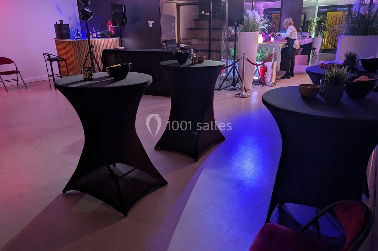 Salle de réception avec tables hautes noires, éclairage coloré et bar en arrière-plan.