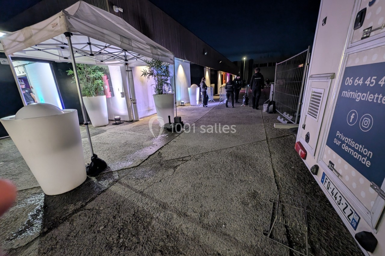 Entrée d'un bâtiment éclairée de nuit avec des tentes blanches, des plantes et des personnes entrant.