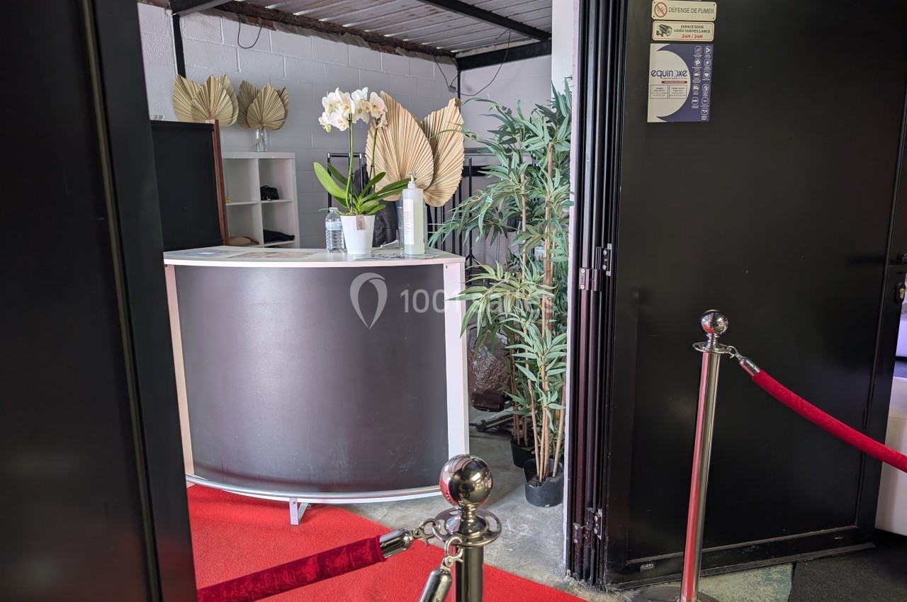 Comptoir d'accueil noir avec des orchidées blanches, plantes décoratives et tapis rouge, délimité par des cordons.