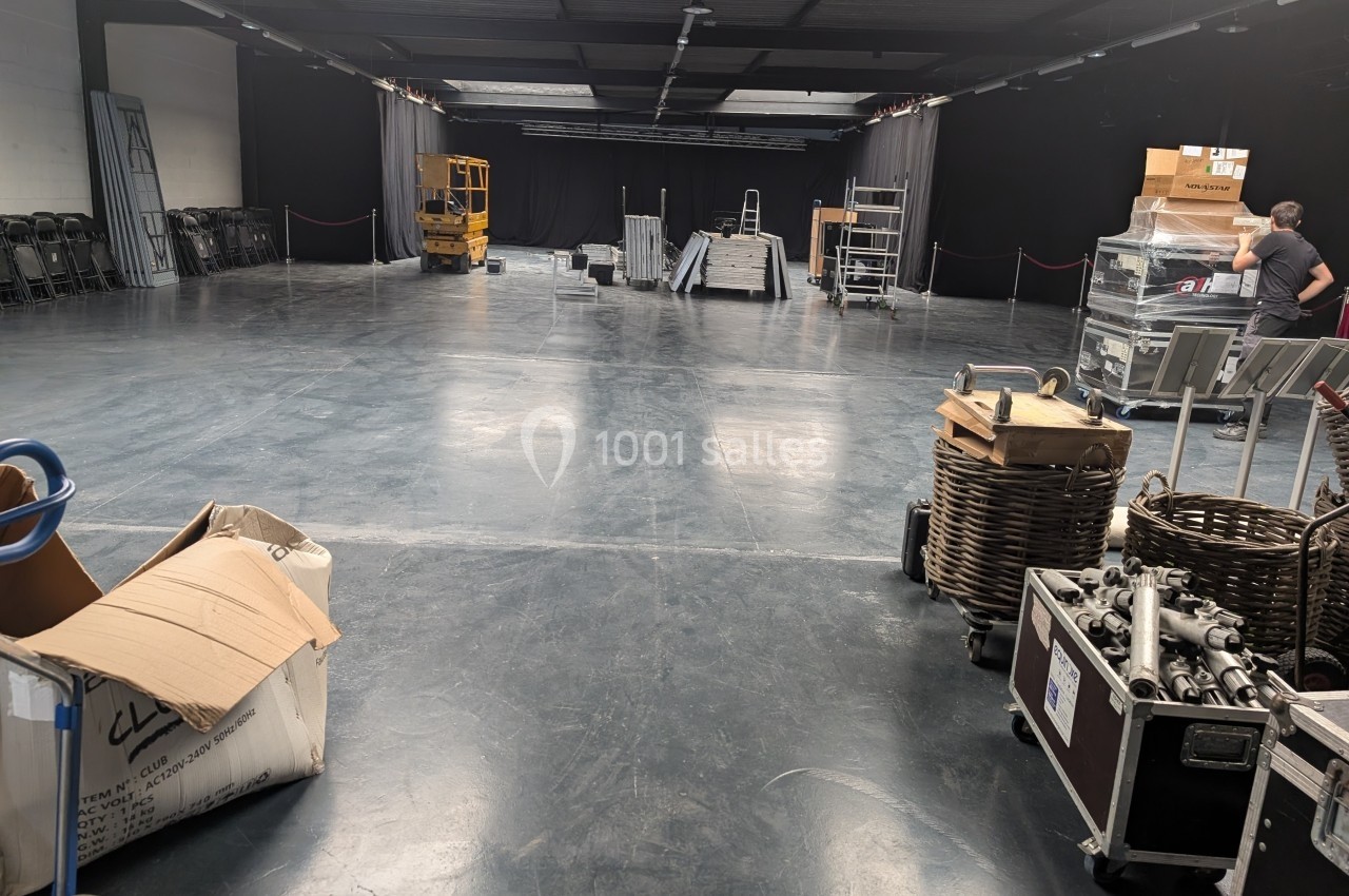 Salle vide en cours de montage avec équipements, cartons, chariots et matériel technique disposés au sol.