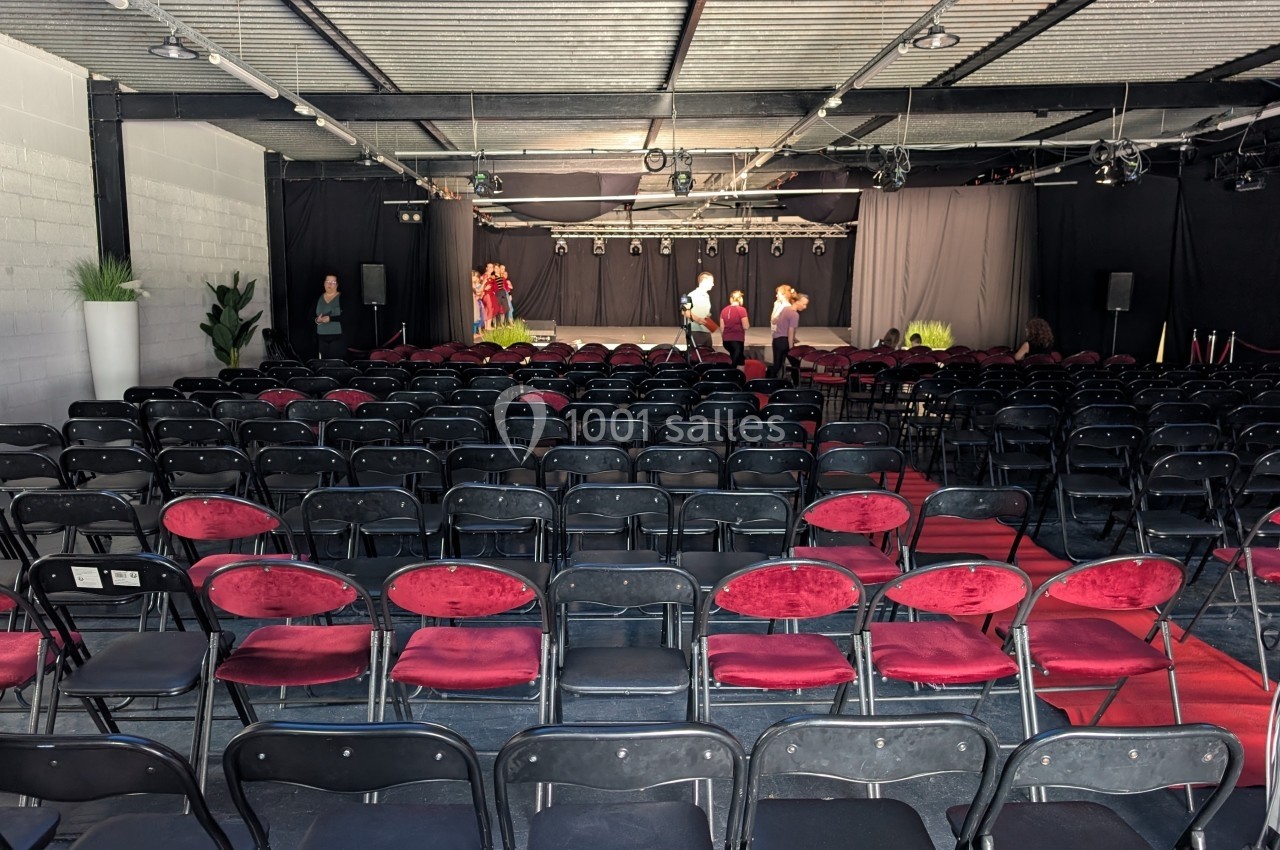 Salle de spectacle avec des rangées de chaises noires et rouges, scène éclairée en arrière-plan et quelques personnes…