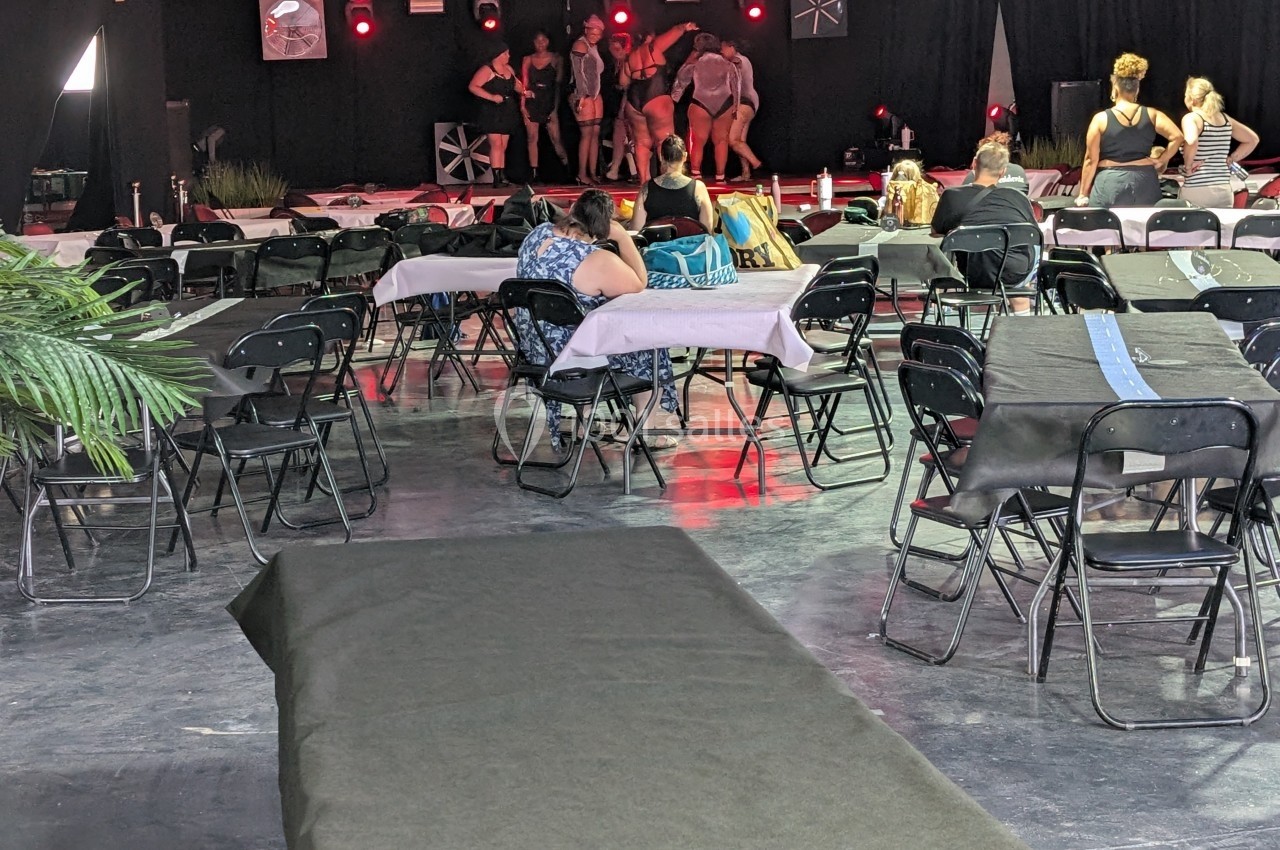 Des personnes assistent à une répétition sur scène dans une salle avec des tables et des chaises disposées autour.