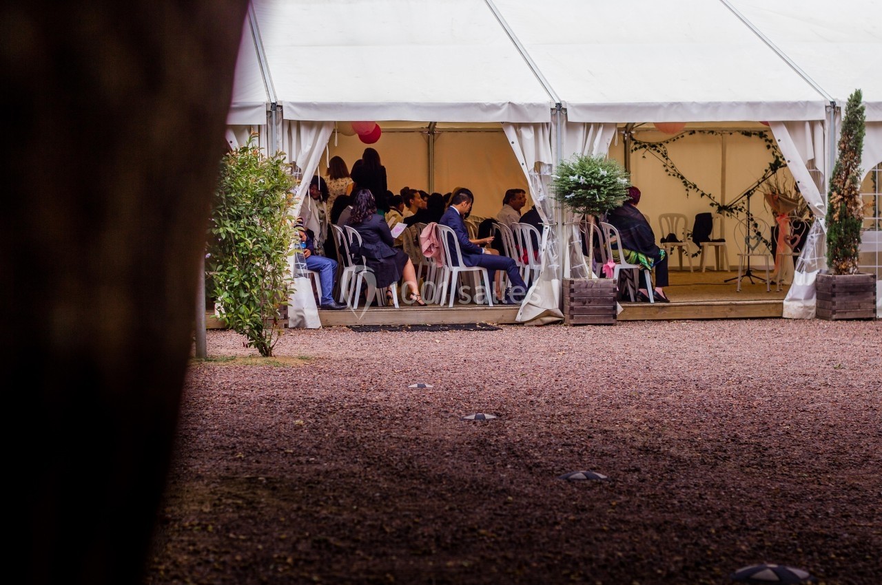 Des personnes assises sous une tente blanche ouverte, entourée de plantes, lors d'un événement en extérieur.