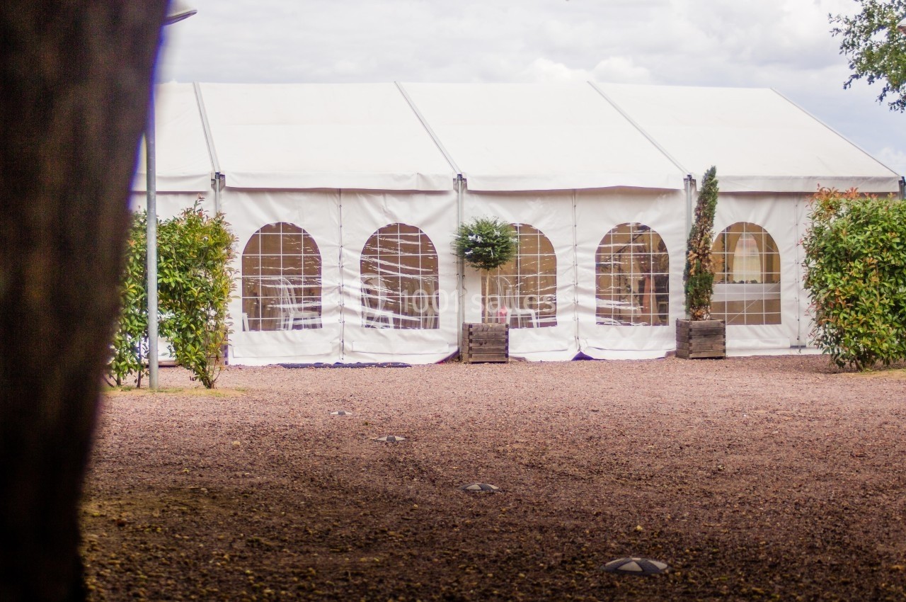 Grande tente blanche avec fenêtres transparentes, installée sur un sol gravillonné entouré de verdure.