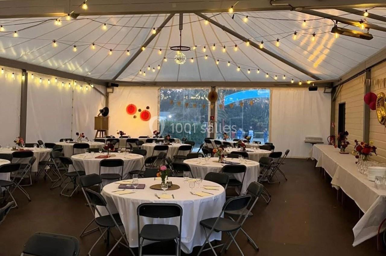 Salle de réception décorée avec des guirlandes lumineuses, des tables rondes dressées et des chaises noires.