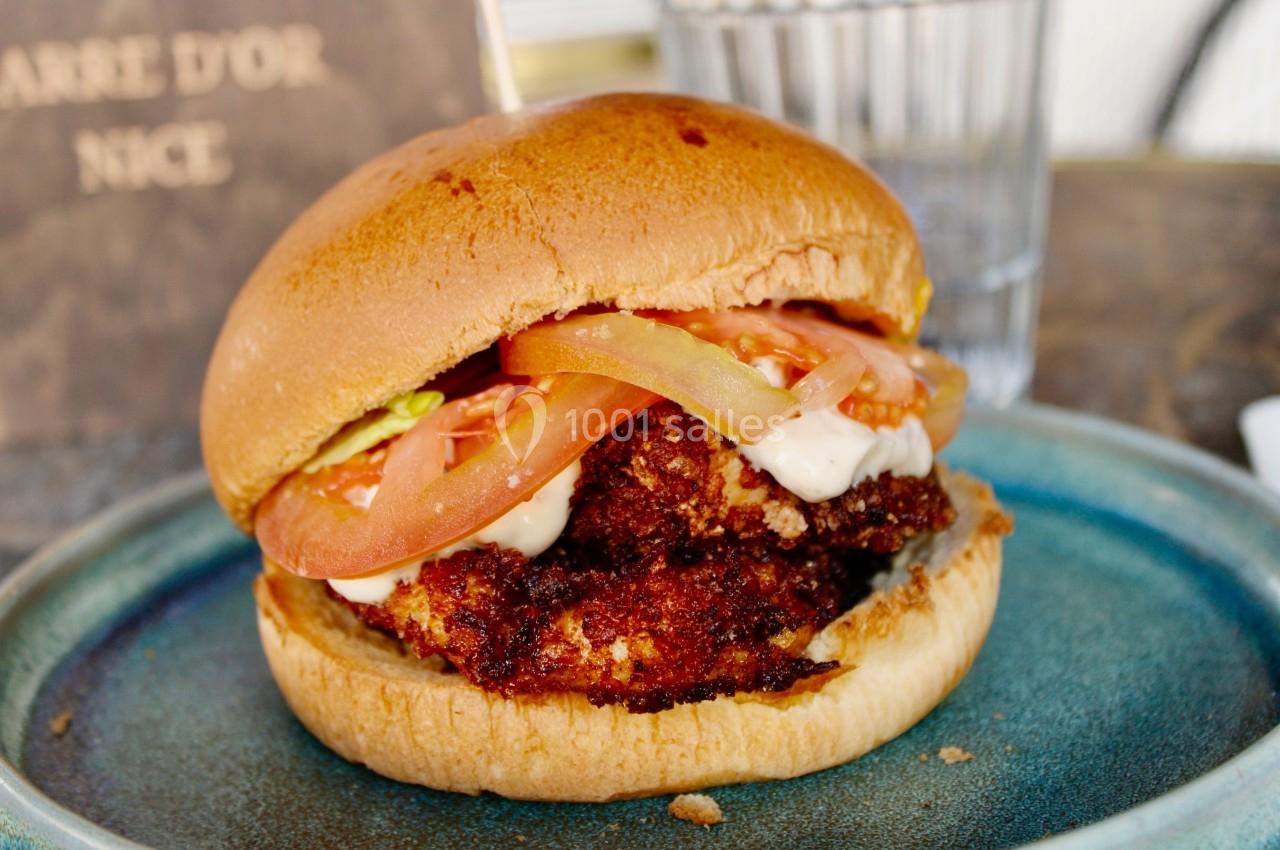 Burger avec pain brioché, poulet pané, tranches de tomate et sauce, servi dans une assiette bleue.