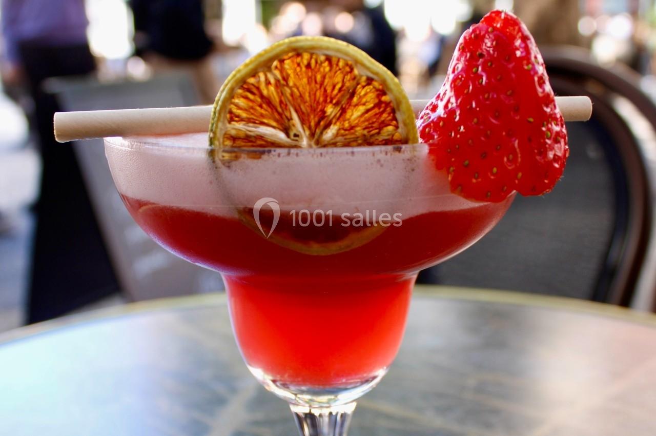 Verre de cocktail rouge garni d'une fraise, d'une tranche d'agrume séchée et d'une paille, posé sur une table.