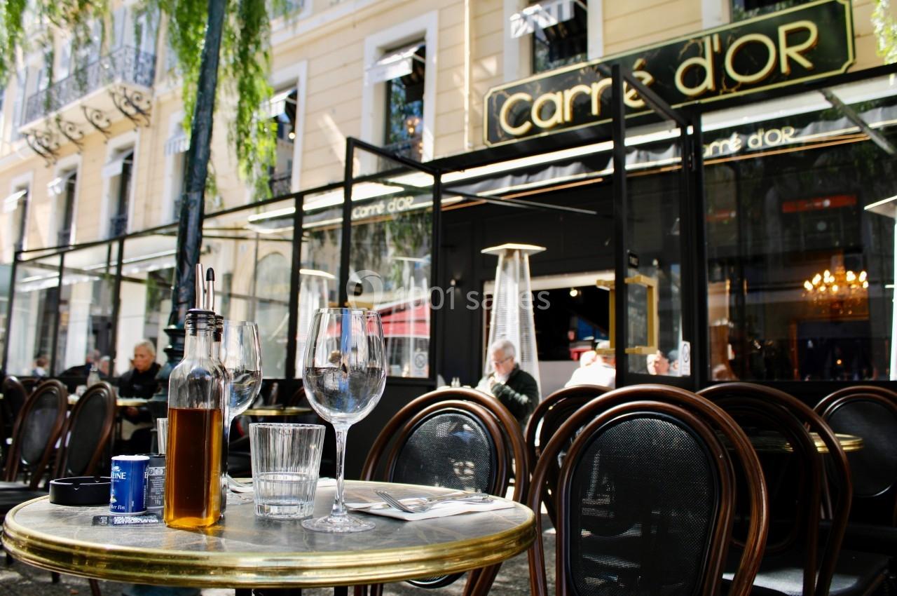 Terrasse d'un café avec des tables en métal, des verres, une bouteille d'huile et des chaises en bois devant une façade…