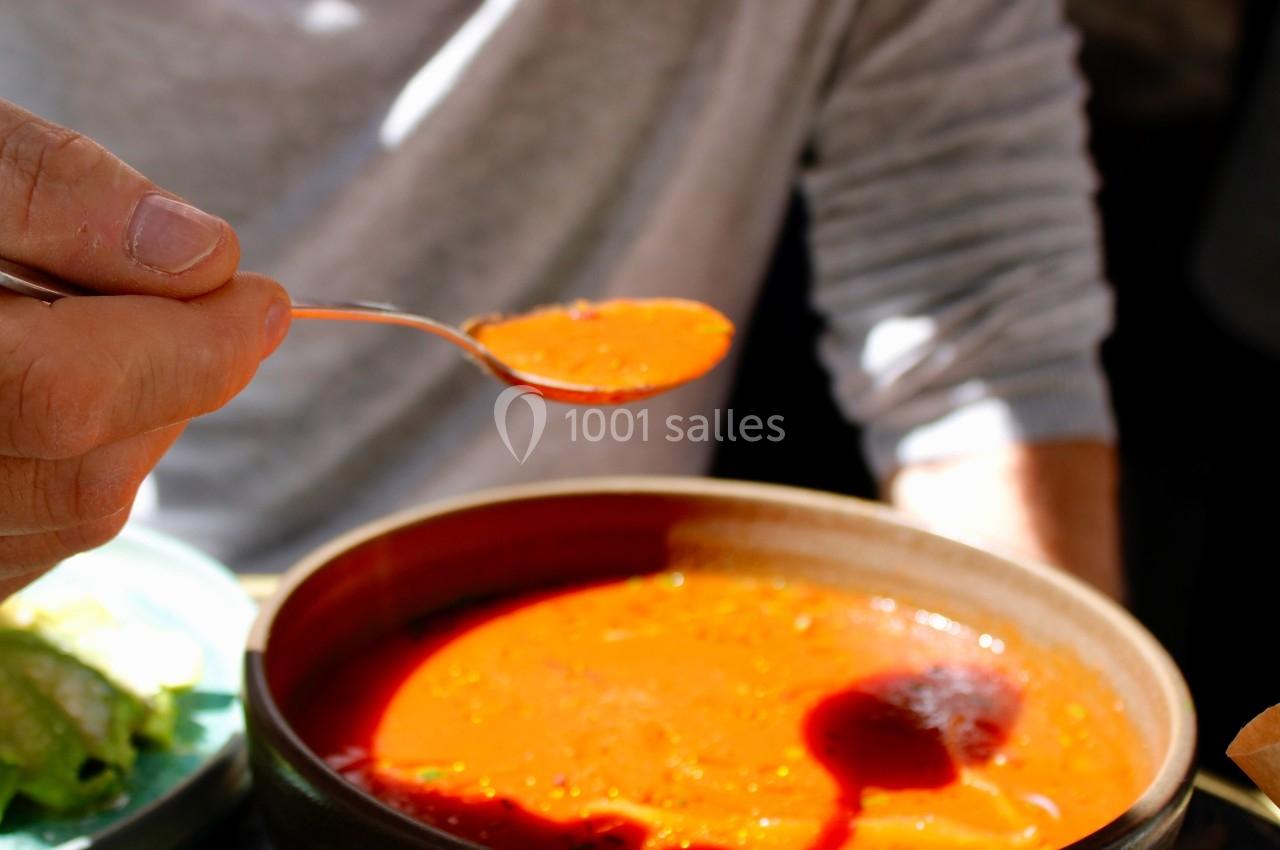 Une personne tient une cuillère remplie de soupe rouge au-dessus d'un bol, éclairée par une lumière naturelle.