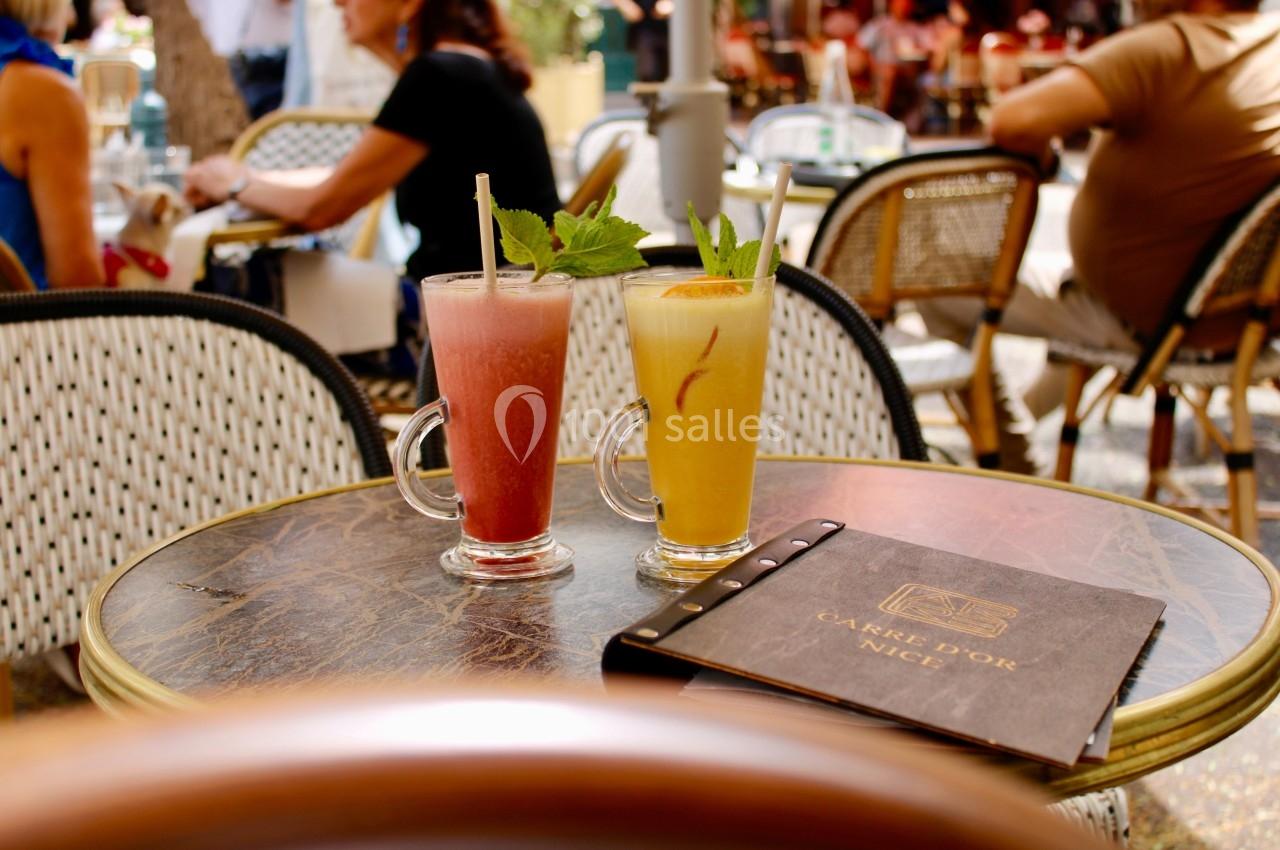 Deux verres de cocktails colorés sur une table en terrasse, entourée de chaises en rotin et de clients en arrière-plan.