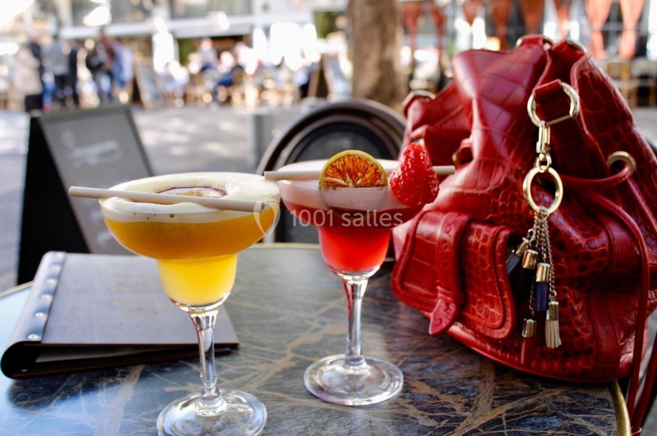 Deux cocktails colorés sur une table en terrasse, accompagnés d'un sac rouge et d'un menu en arrière-plan.