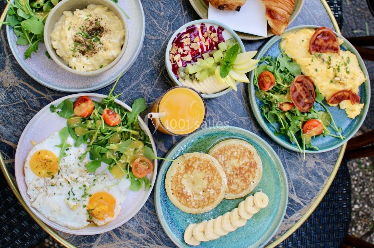 Assortiment de plats pour un brunch, incluant œufs, pancakes, fruits, salade et jus d'orange sur une table en marbre.