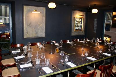 Salle de réunion moderne avec tables disposées en U, chaises en bois, éclairage suspendu et écran mural.