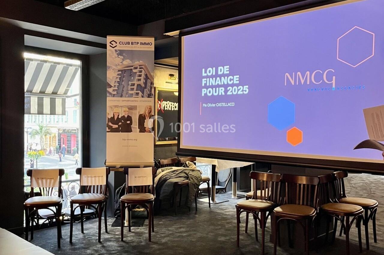 Salle de conférence avec écran affichant ’Loi de finance pour 2025’, chaises alignées et table avec micros et tasses.