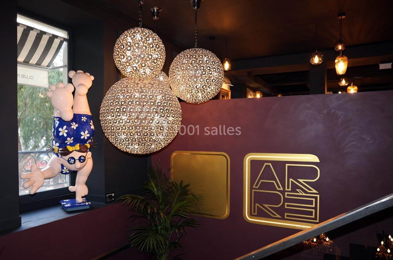 Sculpture colorée d'un personnage en équilibre près d'une fenêtre, éclairage décoratif et logo doré sur un mur violet.