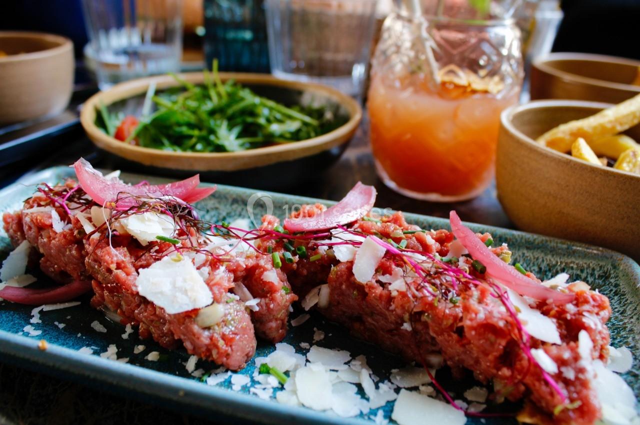 Assiette de tartare de bœuf garni de copeaux de fromage et d'oignons marinés, accompagnée de salade et d'un verre de boisson.