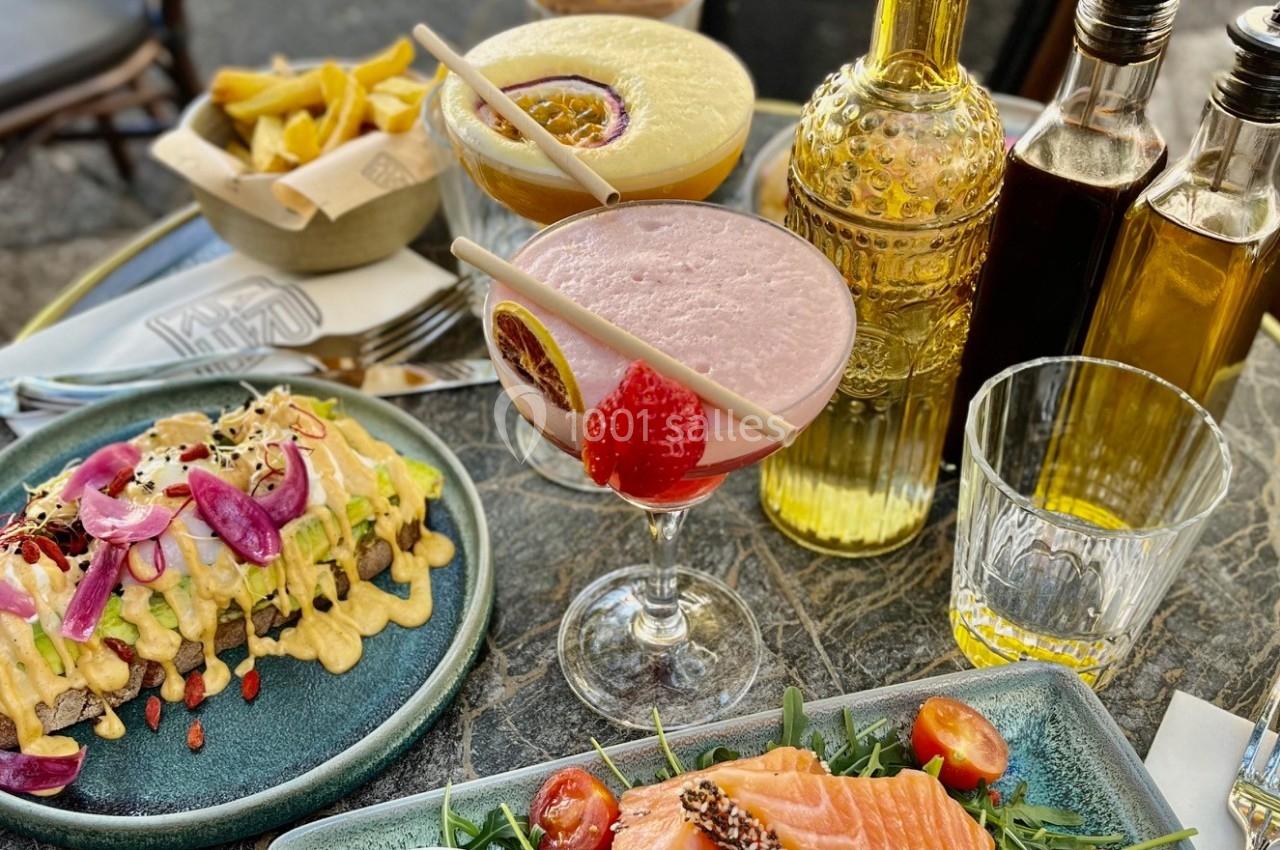 Repas en terrasse avec assiettes de saumon, tacos garnis, cocktails fruités, frites et condiments sur une table en marbre.