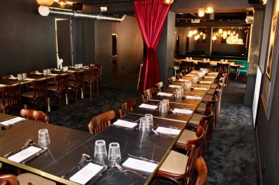 Salle de réunion moderne avec tables disposées en U, chaises en bois, éclairage suspendu et écran mural.