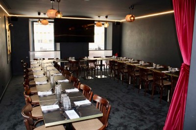 Salle de réunion moderne avec tables disposées en U, chaises en bois, éclairage suspendu et écran mural.