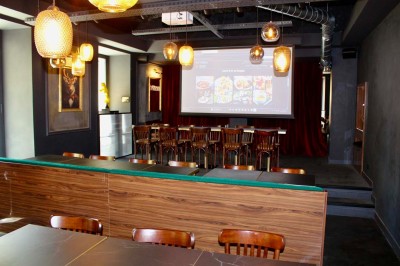 Salle de réunion moderne avec tables disposées en U, chaises en bois, éclairage suspendu et écran mural.