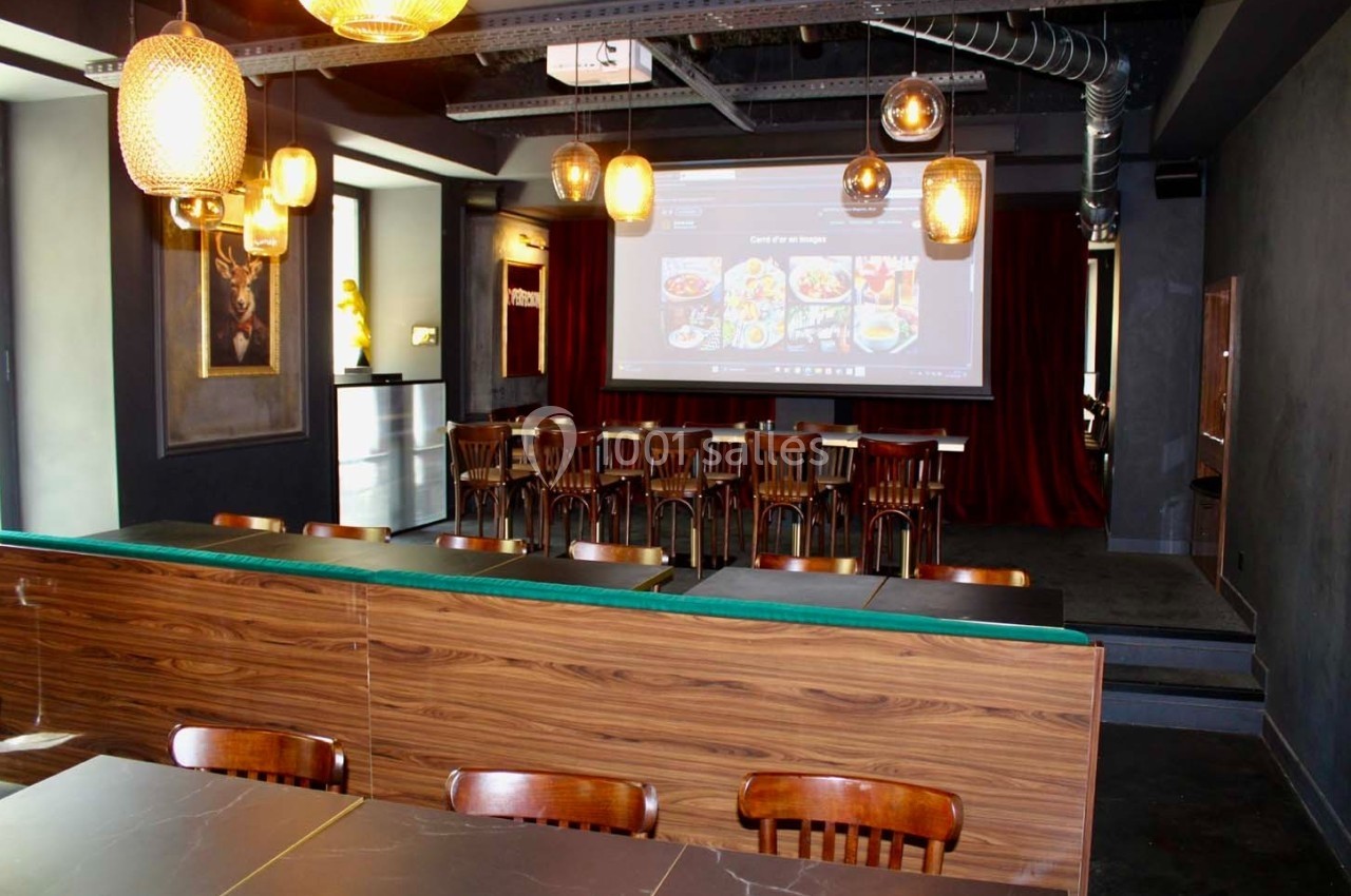 Salle de restaurant avec tables en bois, chaises alignées, éclairage suspendu et écran de projection au fond.