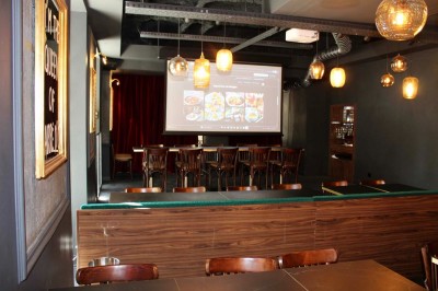 Salle de réunion moderne avec tables disposées en U, chaises en bois, éclairage suspendu et écran mural.