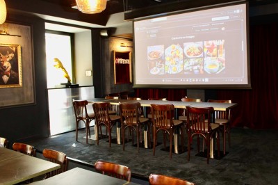 Salle de réunion moderne avec tables disposées en U, chaises en bois, éclairage suspendu et écran mural.