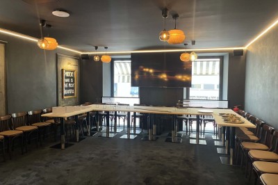 Salle de réunion moderne avec tables disposées en U, chaises en bois, éclairage suspendu et écran mural.