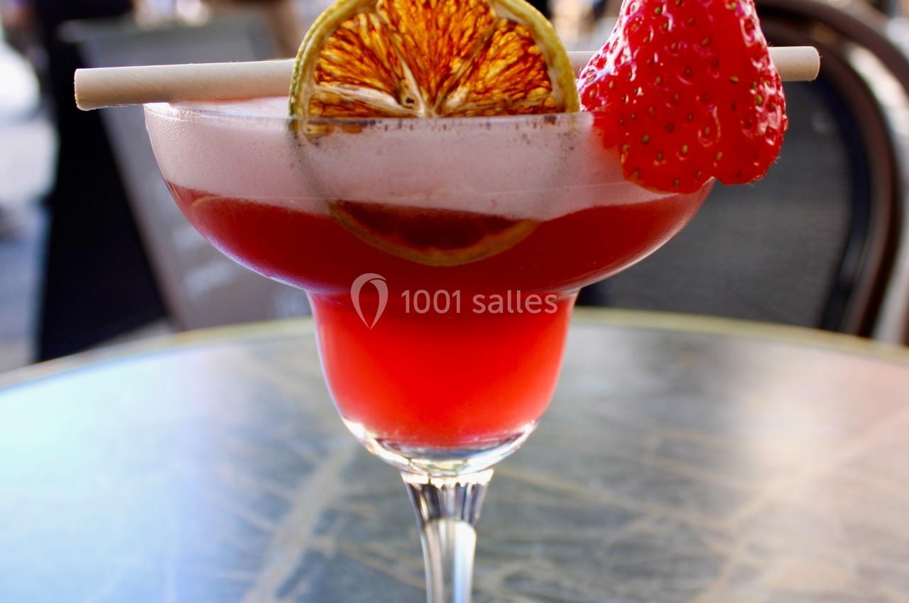 Verre de cocktail rouge garni d'une tranche d'orange séchée et d'une fraise, posé sur une table en extérieur.