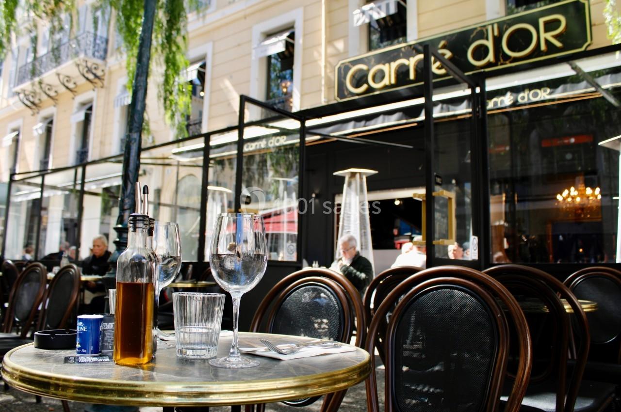 Terrasse d'un café avec des tables et chaises en bois, verres et bouteille d'huile sur une table, devant une façade vitrée.
