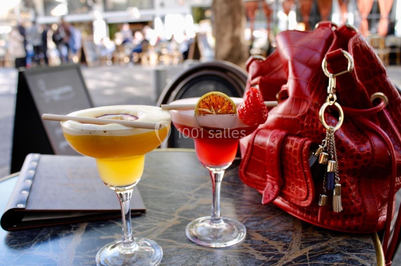 Deux cocktails colorés sur une table en terrasse, accompagnés d'un sac rouge et d'un menu en arrière-plan.