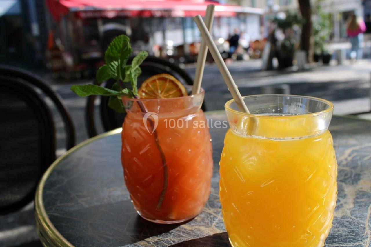 Deux verres de jus colorés sur une table en terrasse, l'un décoré de menthe et d'une tranche d'orange séchée.