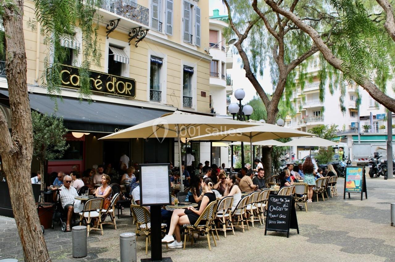 Terrasse animée d'un café en plein air avec des clients assis sous des parasols, entourée d'arbres et de bâtiments.