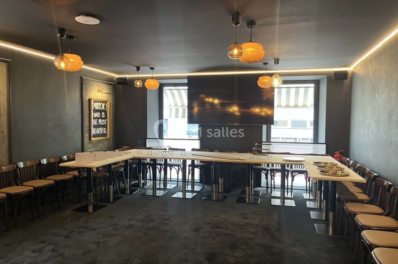 Salle de réunion moderne avec tables disposées en U, chaises en bois, éclairage suspendu et écran mural.