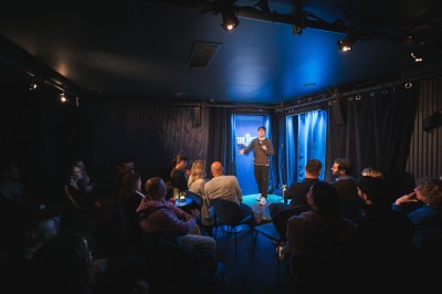 Scène de stand-up vide avec un tabouret, un micro sur pied et un éclairage bleu dans une petite salle intimiste.
