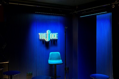 Scène de stand-up vide avec un tabouret, un micro sur pied et un éclairage bleu dans une petite salle intimiste.