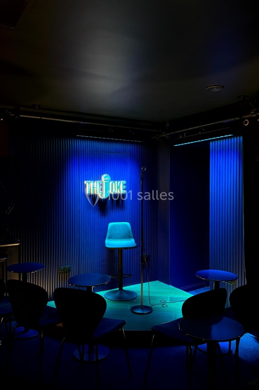 Scène de stand-up éclairée en bleu avec un tabouret, un micro sur pied et des tables dans une salle sombre.