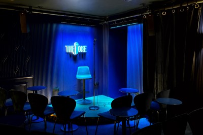 Scène de stand-up vide avec un tabouret, un micro sur pied et un éclairage bleu dans une petite salle intimiste.