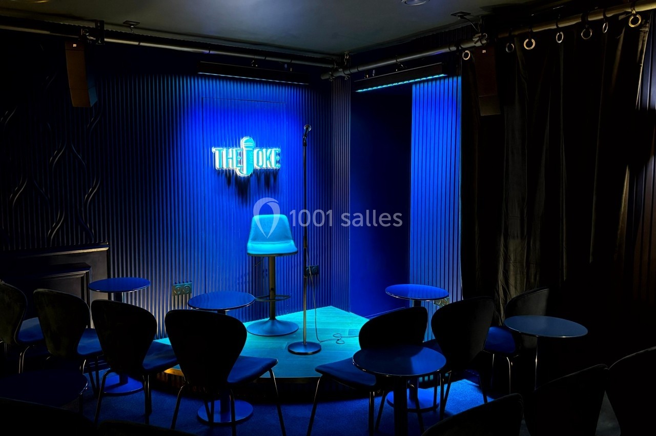 Scène de stand-up vide avec un tabouret, un micro sur pied et un éclairage bleu dans une petite salle intimiste.