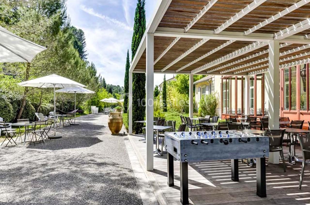 Terrasse ombragée avec tables, chaises, baby-foot et parasols, entourée de verdure sous un ciel dégagé.